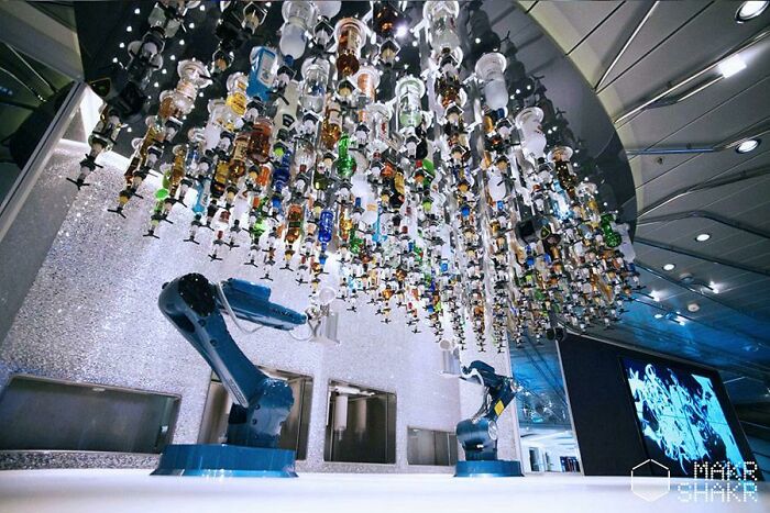 Robotic arms in a futuristic bar, creating unique drinks for a tourist experience with hanging bottles overhead.