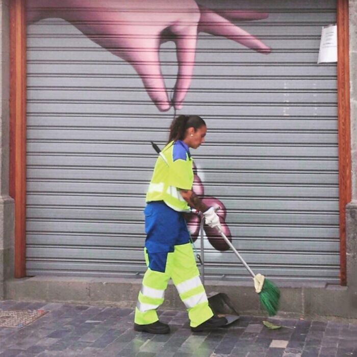 A woman in bright uniform sweeps the street in front of a mural, capturing the extraordinary within the ordinary.