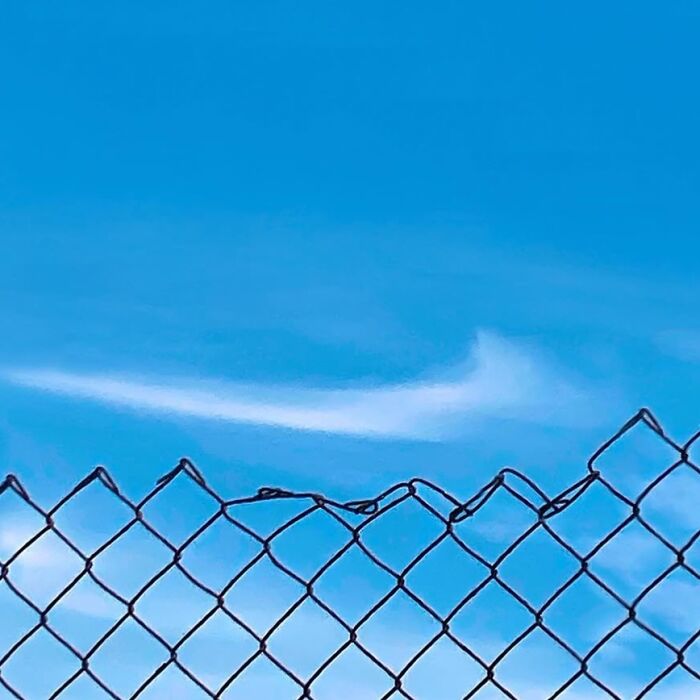 Chain-link fence with a wavy cloud pattern in the blue sky, showcasing the extraordinary within the ordinary.