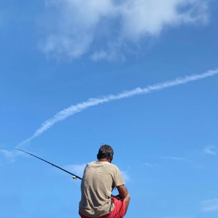 Man fishing under a vast blue sky with wispy clouds, exemplifying Ruiz González’s talent for capturing extraordinary within the ordinary.