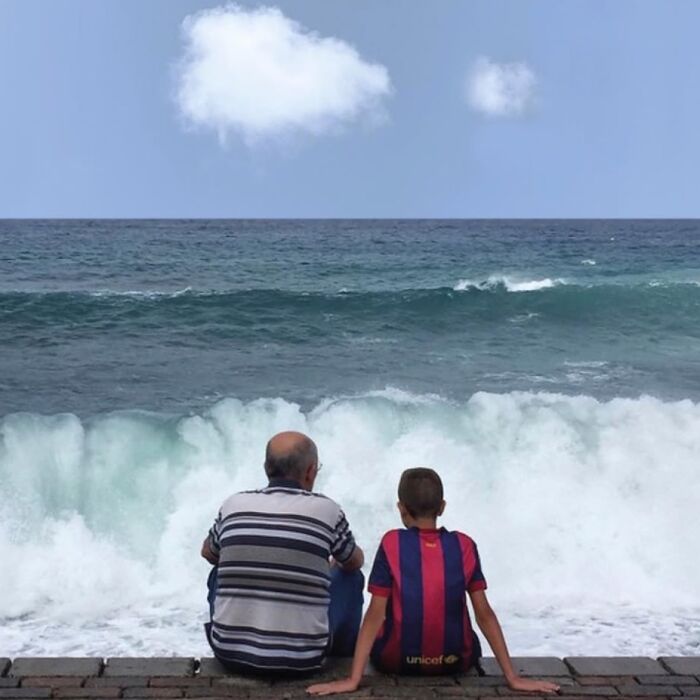 Man and boy seated by ocean waves, clouds above, capturing extraordinary in the ordinary.