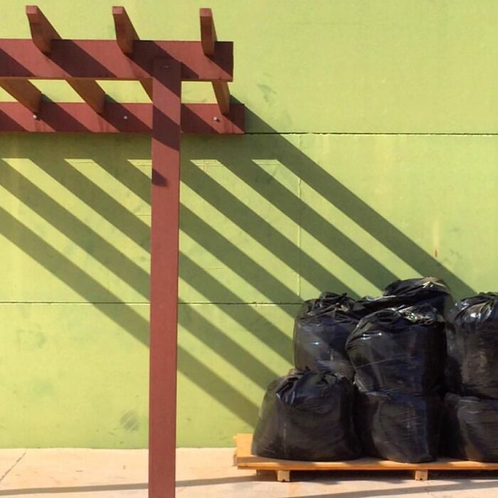 Shadows cast by a wooden structure on a green wall, with black bags piled on a wooden pallet, exemplifying extraordinary within the ordinary.