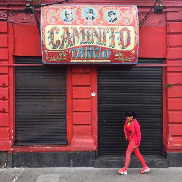 Woman in red walking past a red building with a "Caminito Tango" sign, capturing ordinary moments beautifully.
