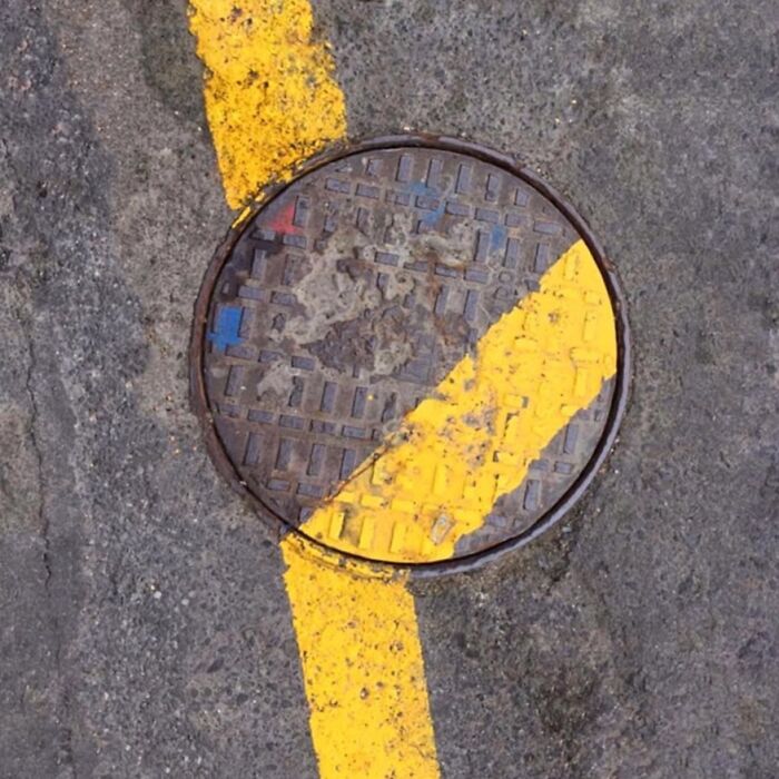 Manhole cover aligned with yellow road stripe, showcasing extraordinary in the ordinary by Jose Ruiz González.