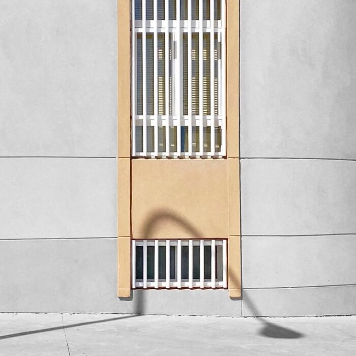 Window with bars against a beige wall, shadow cast below, embodying the extraordinary within the ordinary by Jose Ruiz González.