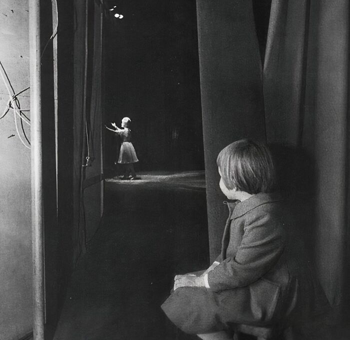 Child in coat backstage, watching a dancer perform under spotlight, capturing true happiness.