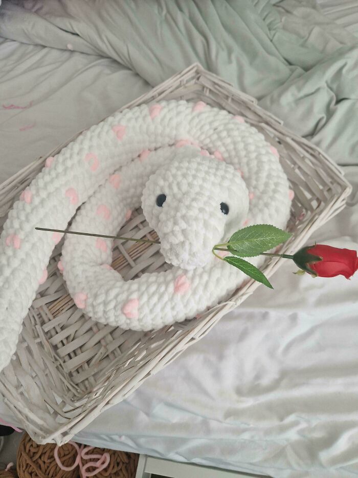 White and pink crocheted snake holding a red rose, displayed in a woven basket on a light green bedspread.