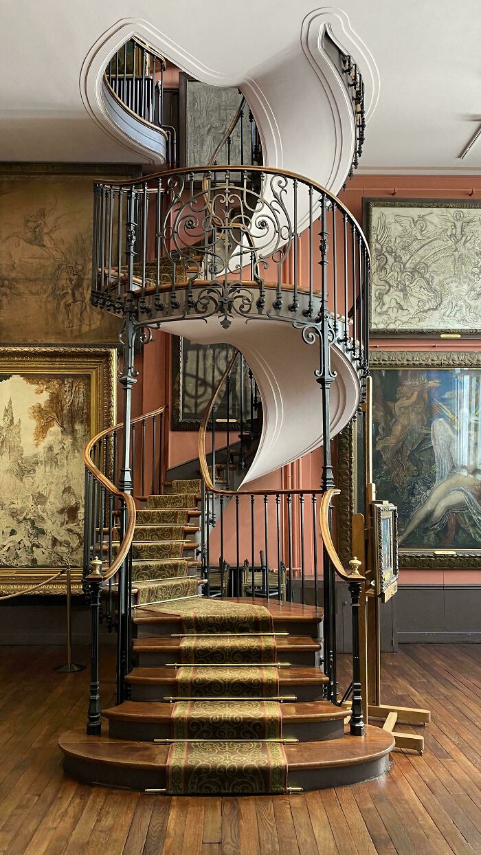 Intricate spiral staircase with ornate iron railings and wooden steps surrounded by impressive architectural feats in a gallery.