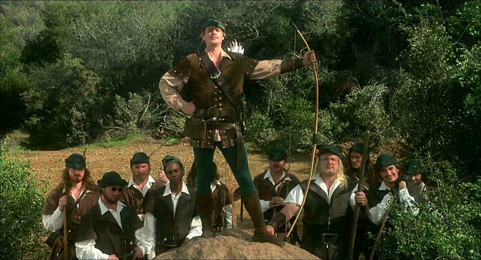 Scene from a '90s movie with a group of men dressed as medieval archers in a forest setting.