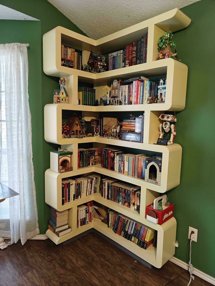 Modern corner bookshelf with artistic woodworker carving details filled with books and miniature models in a cozy room.
