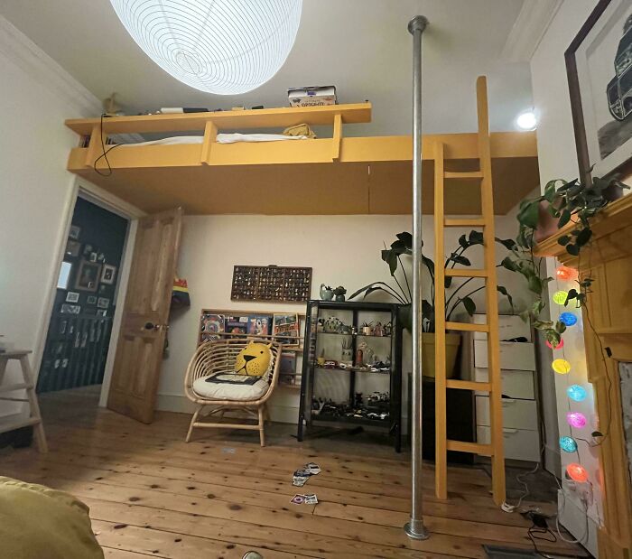 Cozy room with woodworkers' carved loft, wooden ladder, rattan chair, hardwood floor, and decorative shelving with plants.
