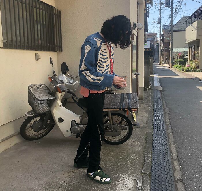 Person wearing a skeleton print jacket, fashion risk, near a parked scooter on a city street.