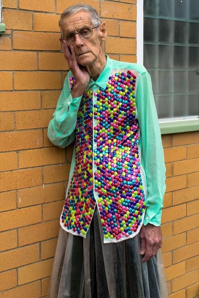 Man showcasing fashion risks with a colorful sequin vest and sheer skirt against a brick wall.