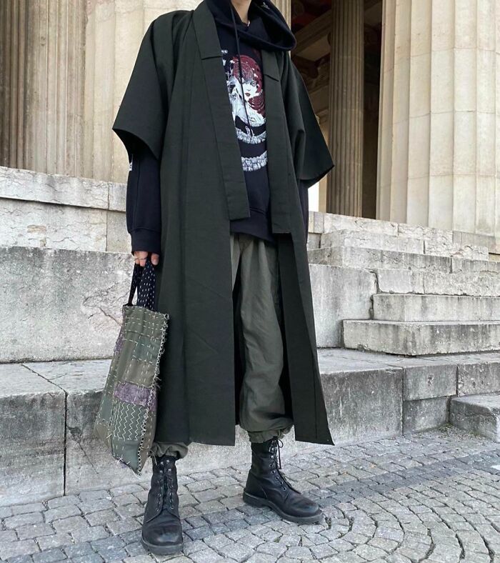 Person taking a fashion risk wearing a long dark coat, graphic hoodie, cargo pants, and boots, holding a textured bag.