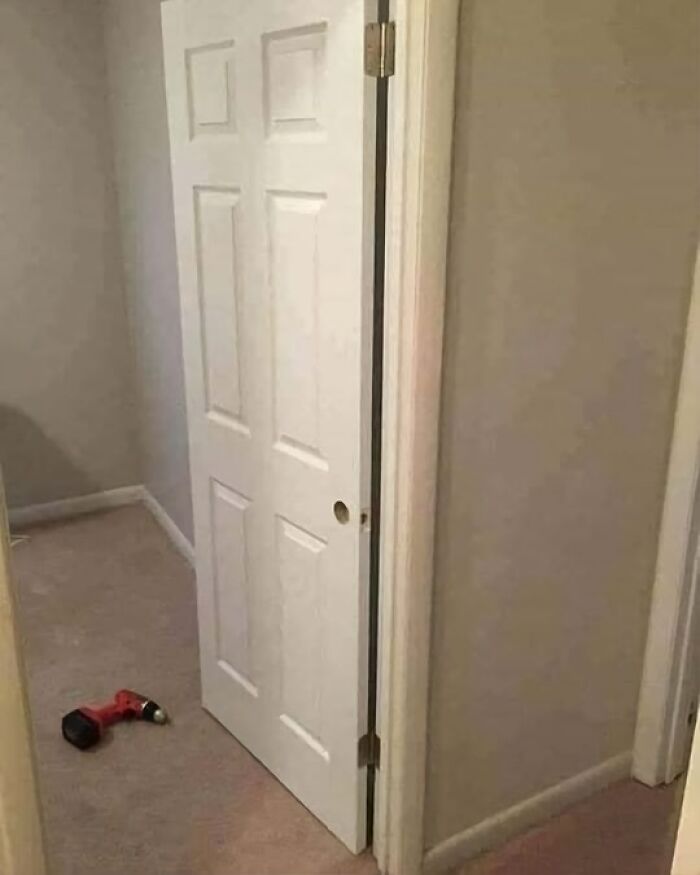 A door installed backward with hinges exposed, showcasing an architecture fail, with a drill lying on the carpet nearby.