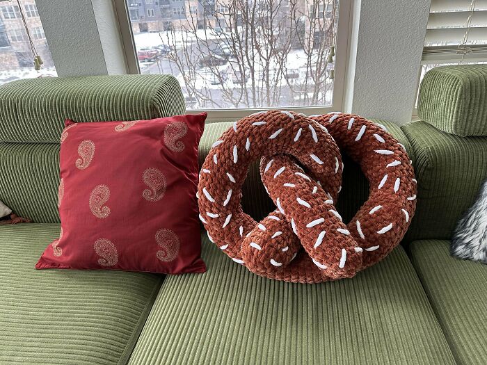 Crochet masterpiece of a plush pretzel pillow with white stitch details on a green couch next to a red cushion.