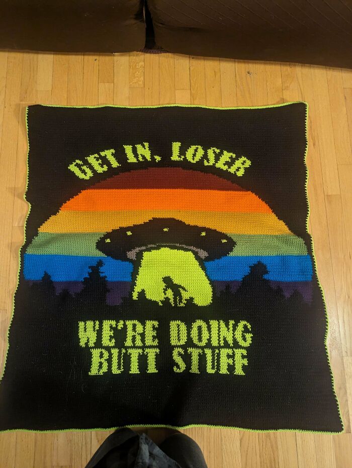 Colorful crochet masterpiece featuring a UFO beam and humorous text on a black background on wooden floor.