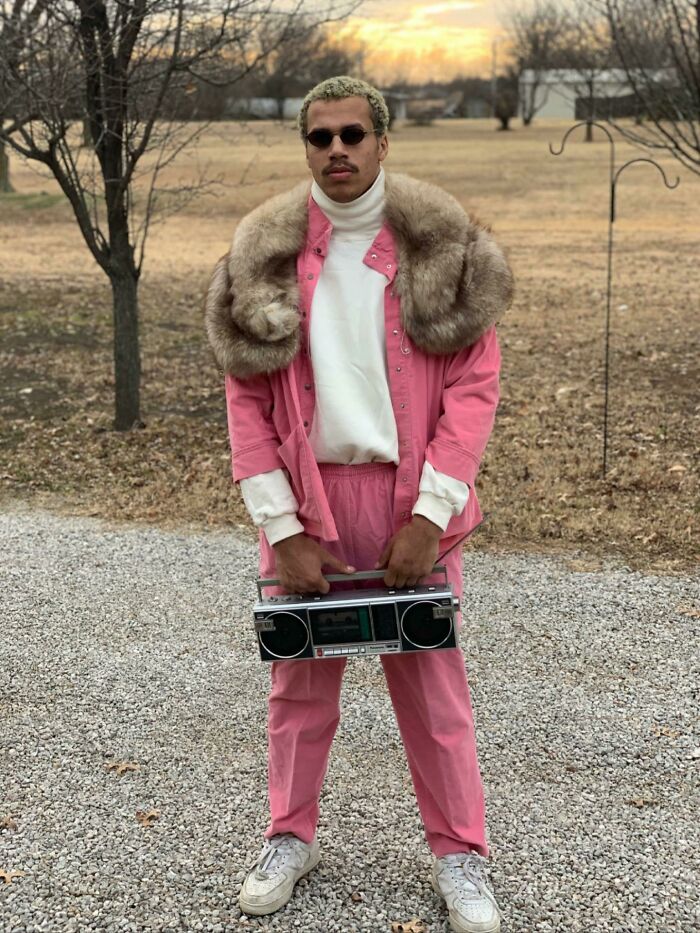 Man in pink suit with fur collar, holding a boombox and wearing sunglasses, showcasing bold fashion risk outdoors.