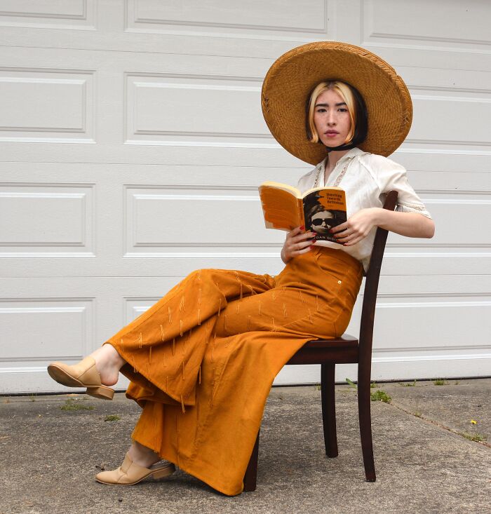 Person taking fashion risks with a large hat and orange pants, sitting on a chair while reading.