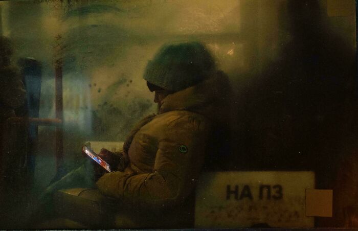 A person in winter attire sits in a dimly lit space, using a phone, reminiscent of an accidental renaissance painting.