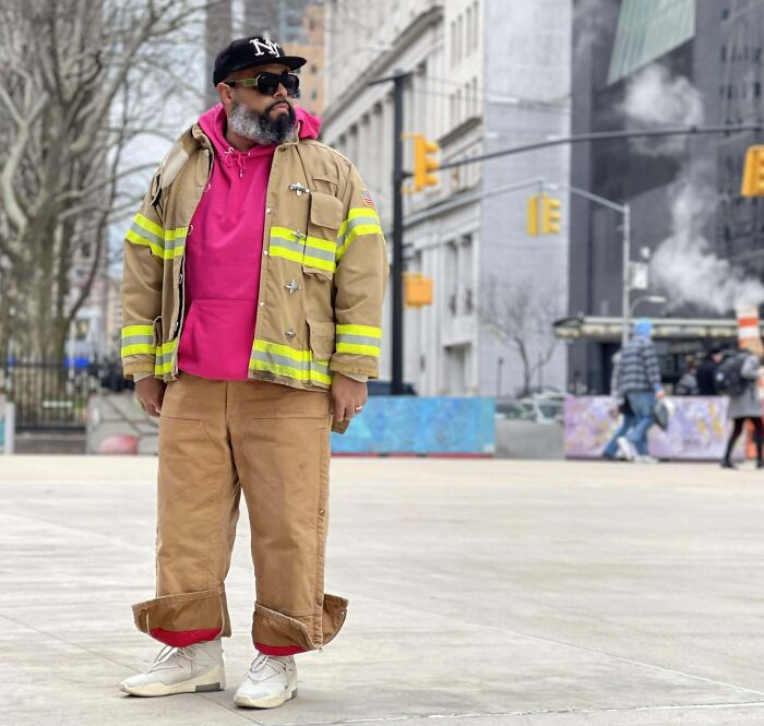 Man wearing a bright pink hoodie and tan firefighter jacket, stylishly showcasing bold fashion risks on a city street.