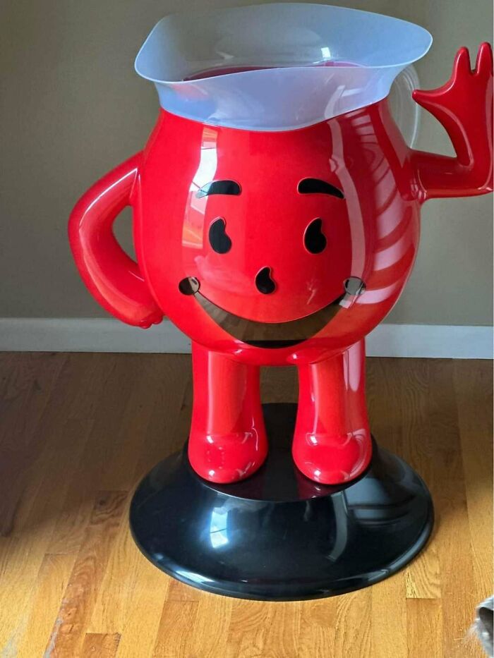 Red vintage toy jug with a smiling face and waving hand, standing on a black base.