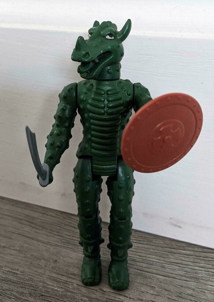 Vintage toy figure, a green dragon warrior holding a sword and shield, standing on a wooden surface.