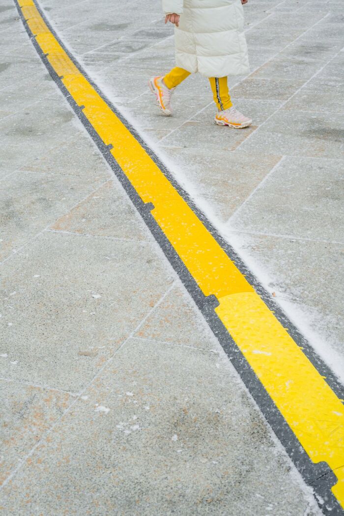 Person in a white coat and yellow pants walking along a snowy street in Oslo and Stockholm, near a yellow line.