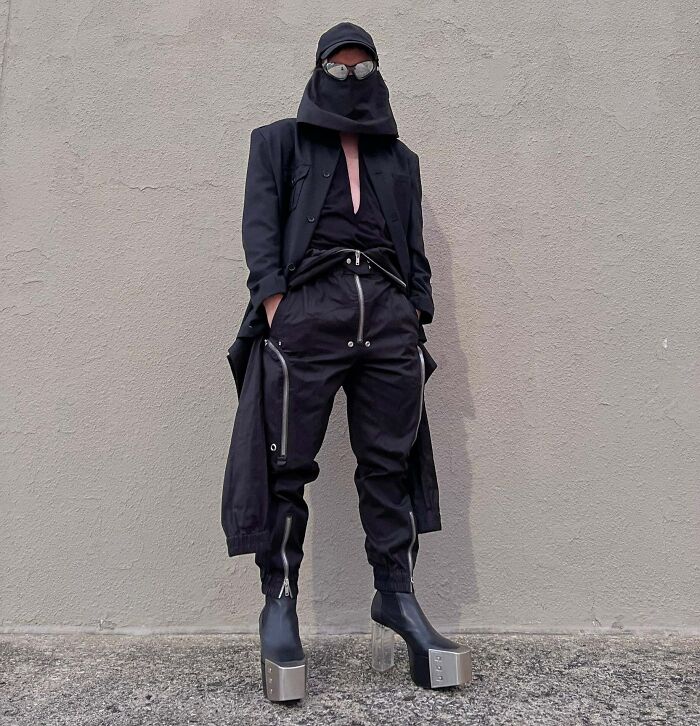 Person taking fashion risks in a unique all-black outfit with high boots and face covering.