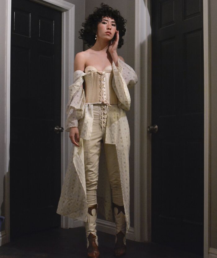 Person taking a fashion risk with a stylish lace-up corset, long sheer coat, and boots in a hallway.