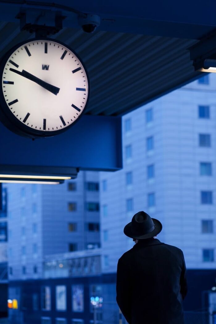 Man in dark coat and hat under large clock, capturing the essence of Oslo and Stockholm streets in a moody, urban scene.
