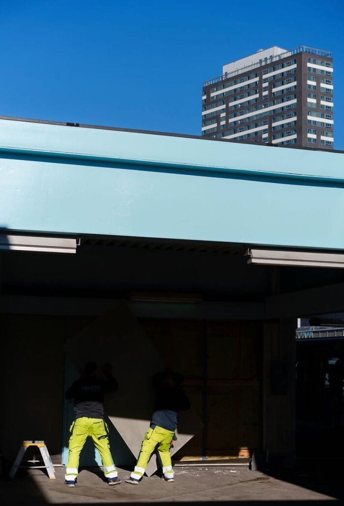 Workers in Oslo's streets, wearing bright gear, handling a large board with a city building in the background.