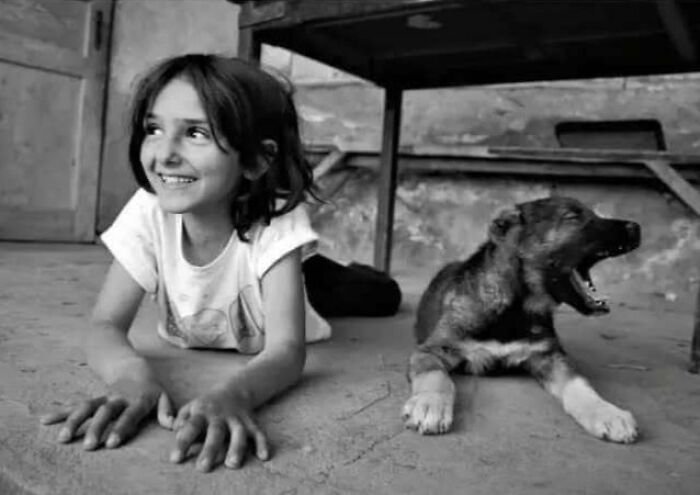 Child smiling on floor next to yawning puppy, black and white photo capturing childhood innocence and resilience.