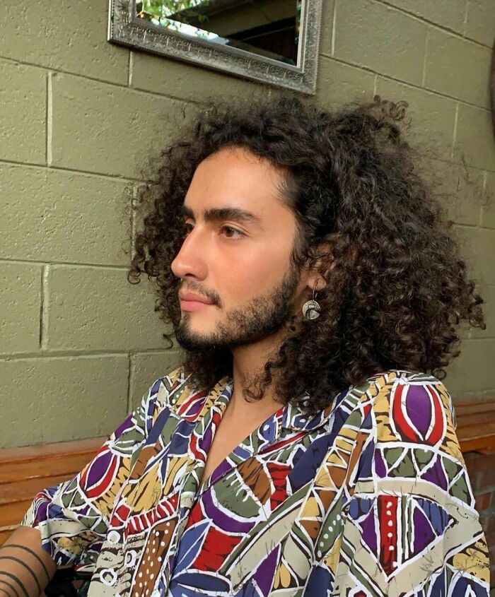 Man with fierceflow beautiful hair and curly locks, wearing a colorful patterned shirt, sitting indoors against a green wall.