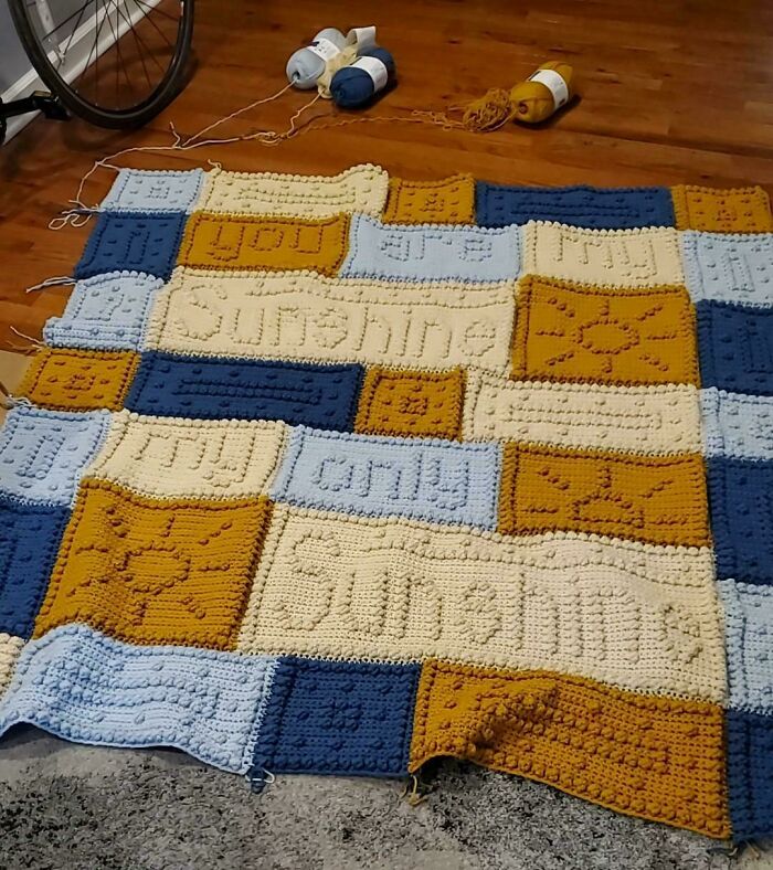 Crochet masterpiece blanket in blue, yellow, and cream with textured sun and text patterns on a wooden floor.