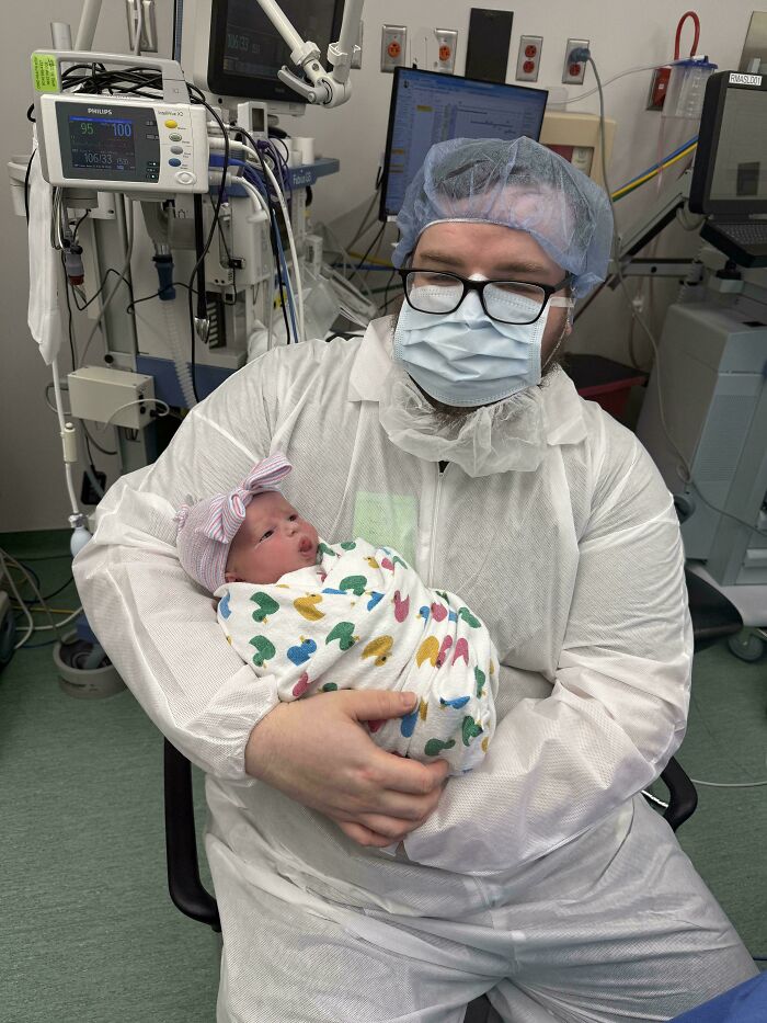 Dad in protective hospital clothing holding newborn baby, showcasing wholesome parenting moments with newborn care.
