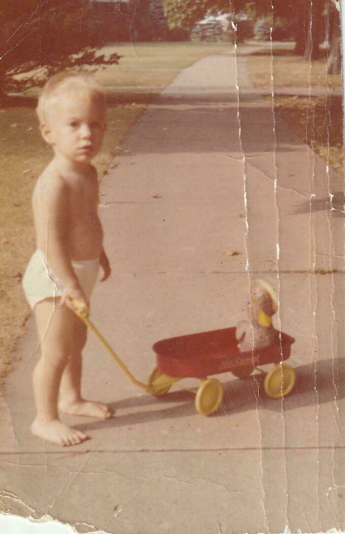 Vintage candid glimpse of a toddler in diaper pulling a small red wagon on a sidewalk in a faded photo.