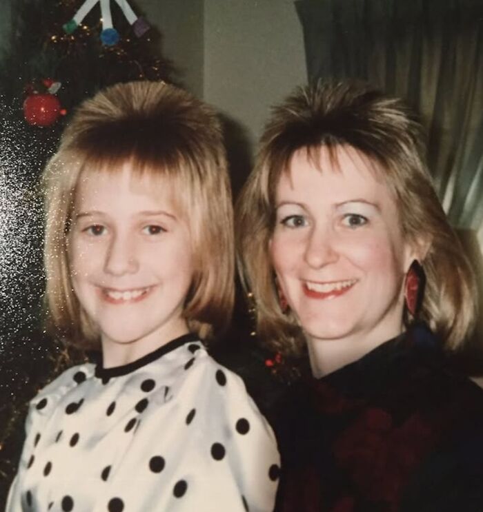Two smiling people with 1980s hairstyles standing together indoors near a decorated Christmas tree.