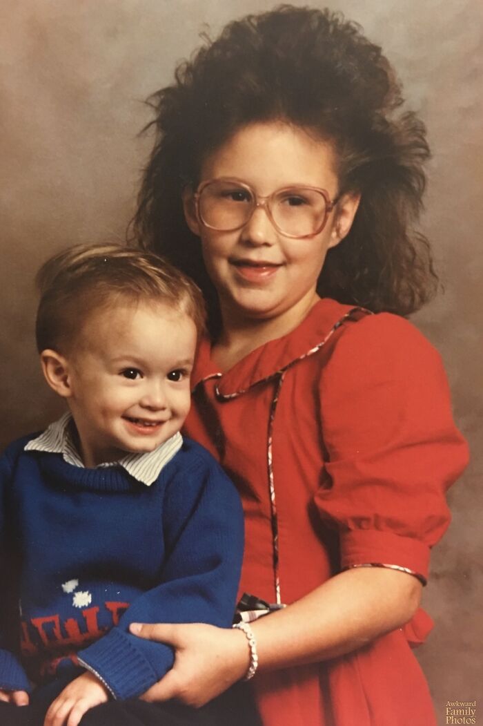 80s hair disaster: Young girl with big teased hair and glasses in red, holding a smiling baby in a blue sweater.