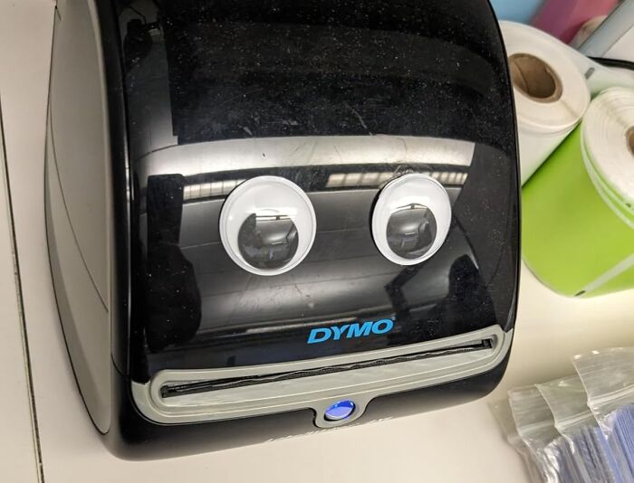 Label printer with googly eyes, resembling an Archie McPhee item, on a desk with label rolls nearby.