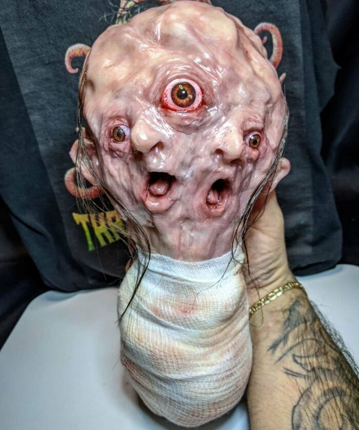 Creepy handmade craft depicting a multi-eyed, distorted face sculpture with human features, held in a tattooed hand.