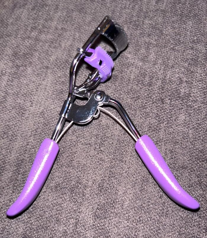 Purple-handled clever invention eyelash curler on a gray surface.