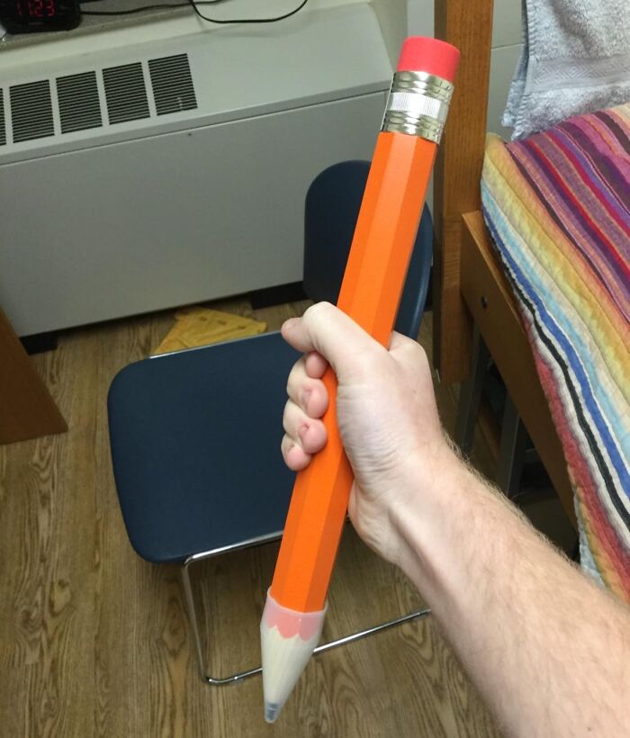 Hand holding a giant novelty pencil, an Archie McPhee item, in a room with a chair and bed.