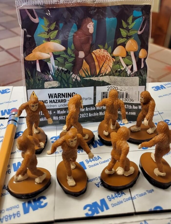 Small Archie McPhee figurines resembling Bigfoot, set against a forest-themed backdrop with mushrooms.