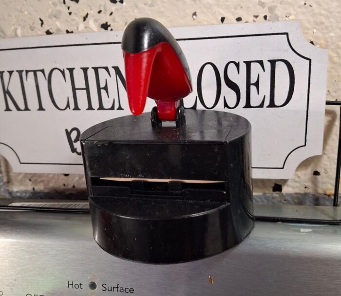 Black and red Archie Mcphee item resembling a bird, sitting on a kitchen appliance near a "Kitchen Closed" sign.