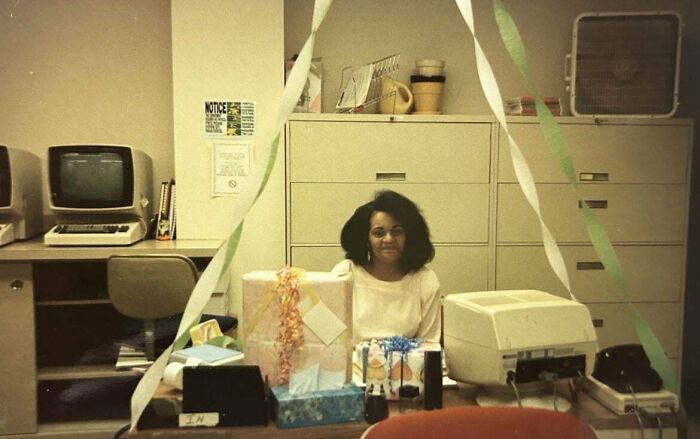Woman sitting at a desk surrounded by vintage computers and wrapped gifts, a candid glimpse into the past office setting.