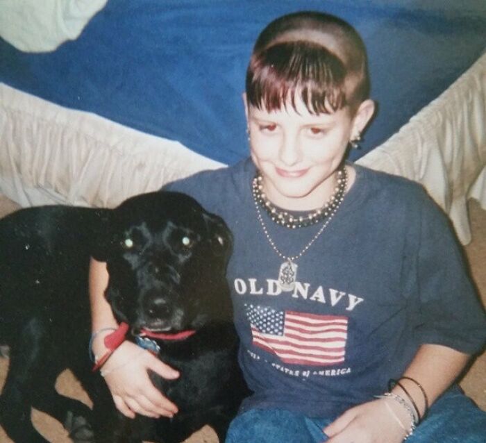 Boy with unique 1990s hairstyle, wearing a flag t-shirt, sitting next to a black dog indoors.