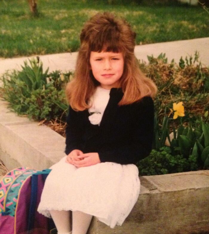 Young girl with voluminous 1980s hairstyle, wearing a white dress and sitting on a sidewalk, surrounded by greenery.