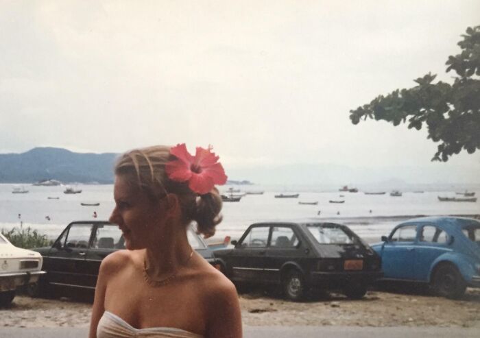 Retro Brazilian beach scene with a woman wearing a flower, vintage cars, and boats on the water.