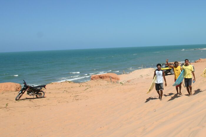 People walking on a sandy Brazilian beach, carrying surfboards under a clear blue sky, with the ocean in the background.